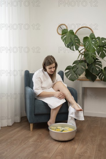 A woman in a white bathrobe enjoys a relaxing foot bath with citrus and flowers at home. The cozy room features minimalist decor, green plants, and a calm, soothing atmosphere for self-care