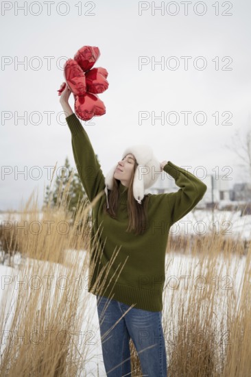 A young woman in a green sweater and white hat lifts red heart shaped balloons in a snowy park. The serene moment captures winter charm and romance