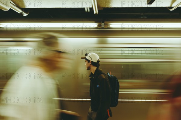 A man pauses in a bustling subway, surrounded by blurred commuters in New York. The scene captures urban solitude, motion, and the daily rush of city life in striking contrast