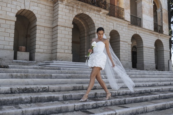 A beautiful bride in a white dress and veil gracefully descends the ancient stone steps of a historic villa in Treia, Macerata, Italy, holding a white flower