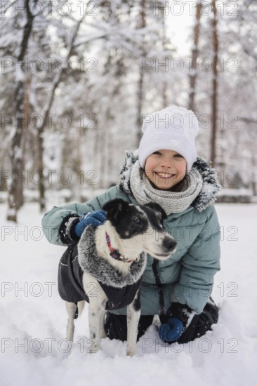 A cheerful child in winter clothing kneels in the snow with a friendly dog. Snow-covered trees surround them, capturing a joyful winter day in the forest