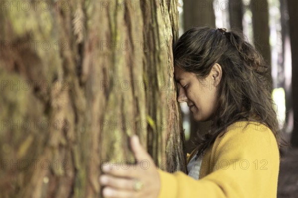 A woman in a yellow sweater hugs a tree in a forest, eyes closed, expressing a deep connection to nature in the Great Ocean Road, Australia. Soft sunlight filters through the trees