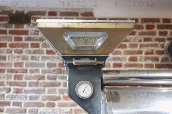 An industrial coffee roasting machine is showcased against a rustic brick wall. The machine holds green coffee beans, preparing for the precise roasting process
