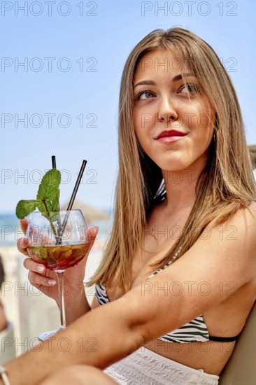 A young woman relaxes at the beach, enjoying a colorful cocktail while soaking up the sun. The waves gently lap at the shore in the background, enhancing the summer vibe