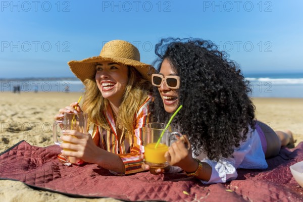 Two cheerful young women lying on a red blanket at the beach, laughing and enjoying their drinks under the bright sun, embracing friendship and a joyful summer holiday experience
