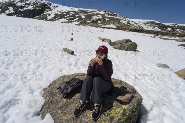 An Asian woman sits comfortably on a rocky surface, enjoying a meal amidst snow-covered peaks in the Sierra de Gredos, Spain. The clear sky adds to the serene ambiance