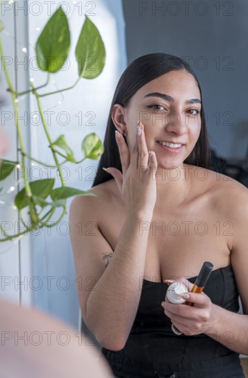 A woman stands by a mirror, applying skincare cream with a smile. Natural light and indoor plants create a calming atmosphere, emphasizing beauty and self care