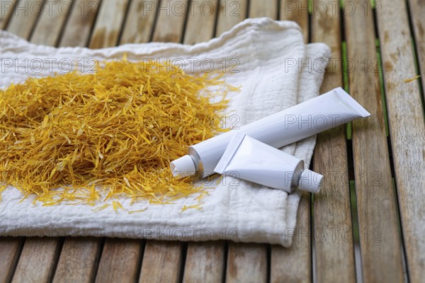 Marigold petals on a cloth and white cosmetic tubes of marigold petals cream on a wooden surface. The image portrays a natural, herbal approach to skincare and beauty products