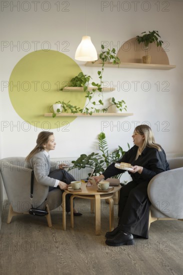 Two women enjoy a friendly chat over coffee in a cozy, stylish cafe with modern decor and lush greenery. Perfect setting for relaxation and meaningful conversation