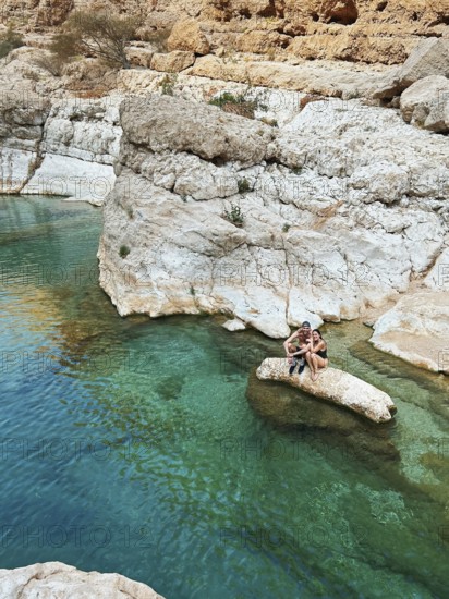 A young smiling couple relaxes on a rocky outcrop in the emerald waters of Wadi Ash Shab, a picturesque oasis located in Oman. This tranquil spot is surrounded by rugged cliffs, enhancing the natural beauty of the area