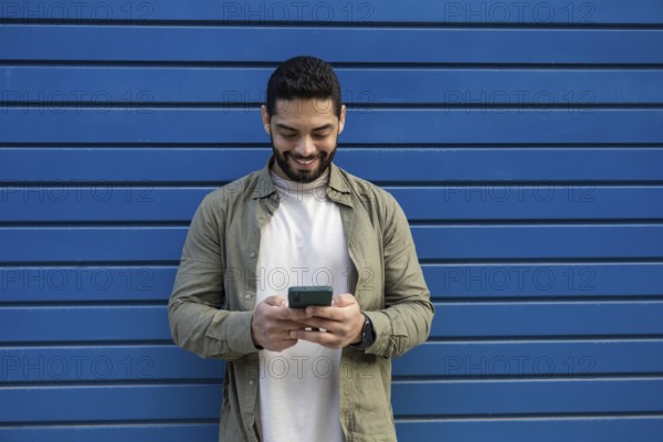 A man in a green shirt and white T shirt smiles while using his smartphone in the city. He stands against a bright blue wall, creating a modern and cheerful atmosphere