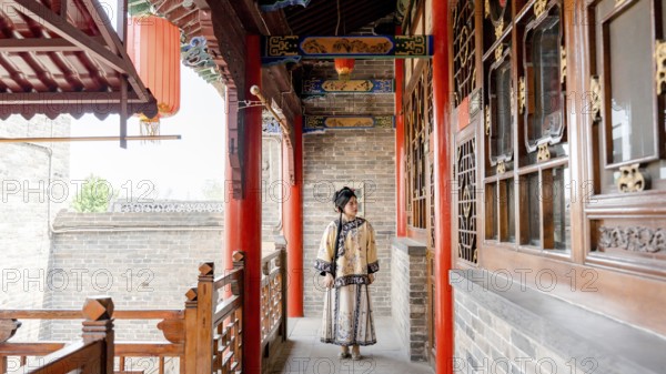 A young woman dressed in traditional Qing Dynasty attire stands gracefully in a historic corridor in Pingyao, China. The setting highlights intricate wooden carvings and classic decor