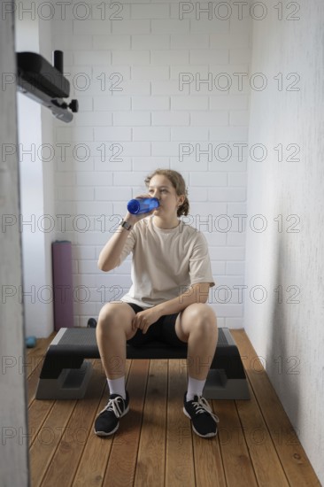 A young person takes a refreshing break with a water bottle in a compact home gym, sitting on a step bench, emphasizing the importance of hydration while exercising