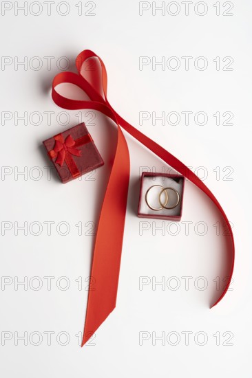 From above, a romantic Valentine's Day setup featuring a red ribbon arranged in a heart shape, with a small red gift box and a ring box containing two gold wedding rings, all on a white background