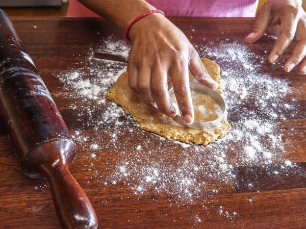 Hands using a circular cutter to shape cookie dough on a floured wooden surface. A rolling pin is next to it, indicating a baking process in a domestic kitchen