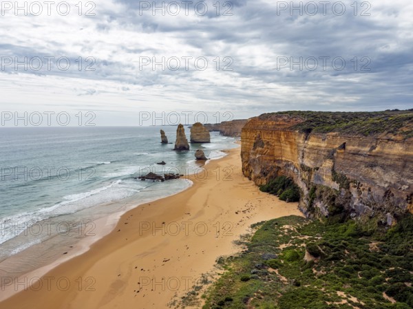 Breathtaking coastal scene featuring towering cliffs and striking sea stacks in the Twelve Apostles, Australia. A sandy beach stretches along the shoreline under a dramatic cloudy sky