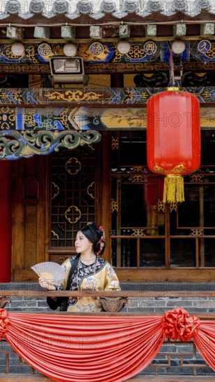A woman dressed in traditional Qing Dynasty attire holds a fan while standing on a decorated balcony in Pingyao, China, under a vibrant red lantern and ornate architecture