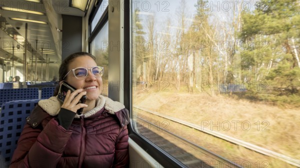 A woman in a cozy jacket is talking on her phone while traveling on a train. She gazes happily out the window at a sunlit landscape, enjoying the scenic journey