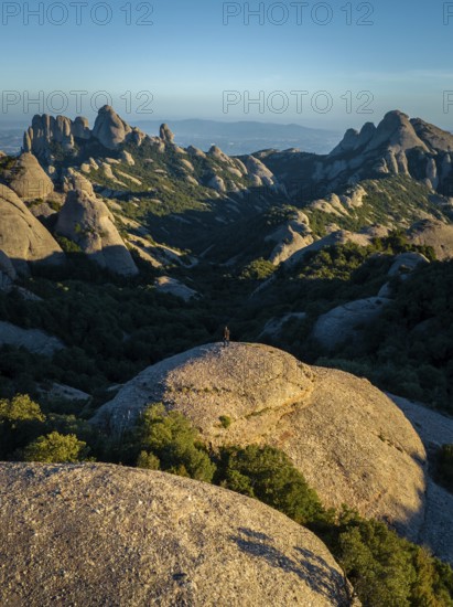 A breathtaking view of Montserrat's rugged rock formations in Catalonia, Spain. The image captures a lone figure standing on a large boulder, surrounded by lush greenery