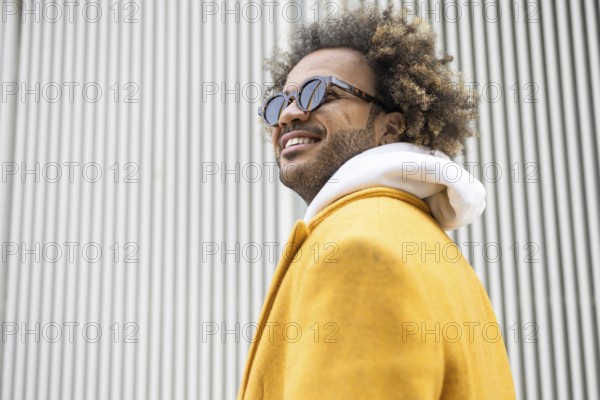 A confident afro man wearing sunglasses and a stylish yellow coat poses in front of a modern striped background, exuding a cheerful and fashionable vibe