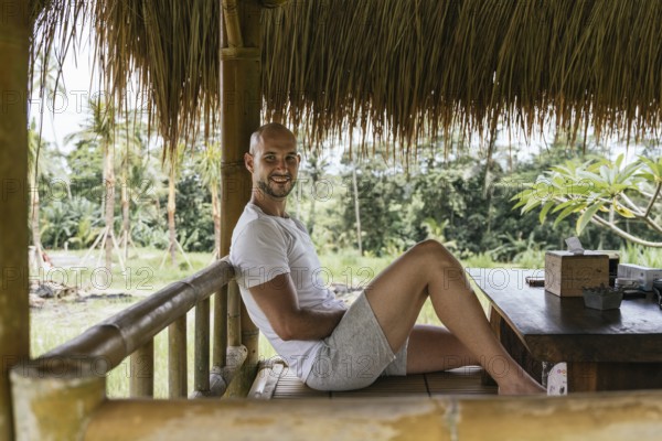 A man in casual attire smiles while relaxing in a bamboo hut, surrounded by lush greenery in Bali. The natural setting and rustic seating create a serene, idyllic atmosphere