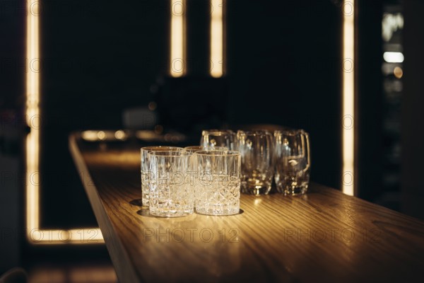 A collection of empty glasses sits on a polished wooden bar, illuminated by warm ambient lighting, evoking a sophisticated and inviting atmosphere