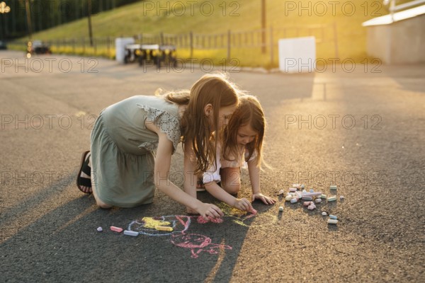 Two kids draw with colorful chalk on asphalt during a bright, sunny day, surrounded by lush greenery. A scene of creativity and childhood joy unfolds in the warm light in summer