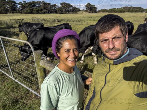 A man and a woman takes a selfie on a sunny day in a grassy field, surrounded by black cows in Australia. The scene captures rural farm life, featuring clear blue skies and vibrant greenery