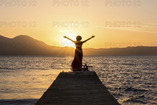 A woman stands on a wooden pier at Papikinou Beach, Milos Island, Greece, embracing the sun as it sets, casting a golden glow over the tranquil sea