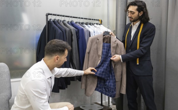 A tailor assists a customer in selecting a suit in modern store. The tailor shows the inner lining of a jacket while the customer examines it. Background features a rack of various suits