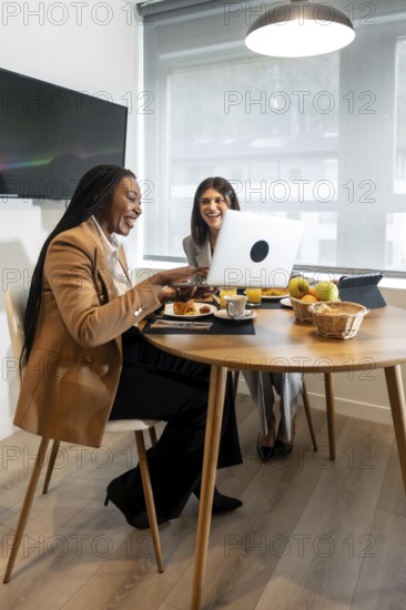 Two businesswomen are enjoying breakfast and working together on a laptop in their hotel room during business trip