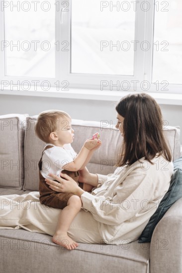 A mother and her son share a tender moment on the couch. The son hands something to the mother, surrounded by a bright, cozy living room setting