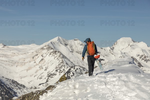 Back view of an unrecognizable person hiking up a snow-covered mountain with a snowboard. The snowboarder, dressed in winter gear, is surrounded by a breathtaking alpine landscape