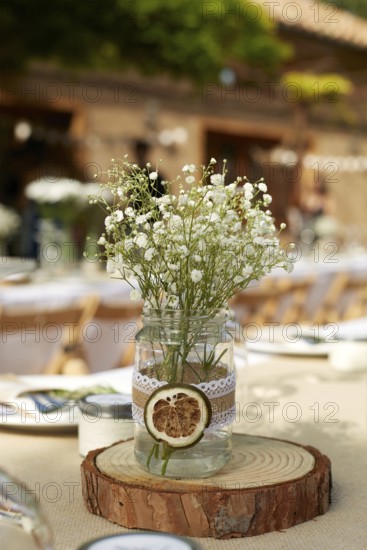 A rustic wedding table centerpiece featuring delicate white flowers in a mason jar adorned with lace, placed on a wooden slab. Ideal for romantic boho-themed weddings
