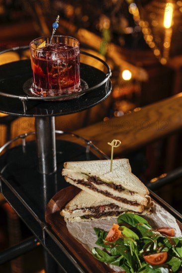 A gourmet club sandwich filled with tender veal cheeks, served alongside a fresh tomato and spinach salad. Accompanied by a classic negroni cocktail for a perfect pairing