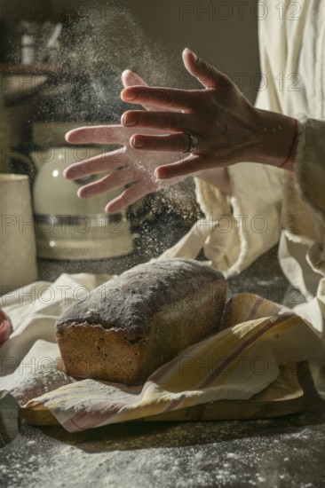 A warm, rustic scene of baking bread. Hands clap to dust flour while a loaf of bread cools on a wooden cutting board, capturing a minimalist image and the essence of homemade bread making