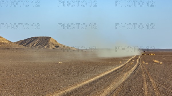 A dusty track runs through a wide, flat desert landscape with hills in the background, with an off-road vehicle through the Sahara desert in Algeria