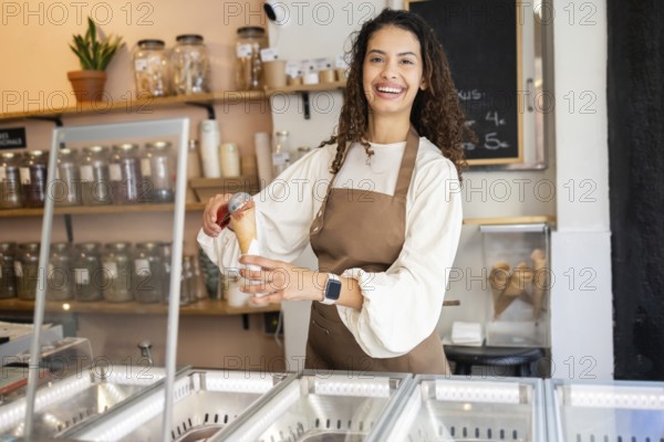 A cheerful woman working in an ice cream shop, serving a scoop of delicious ice cream in a cone. She wears a brown apron and stands behind the counter with joy