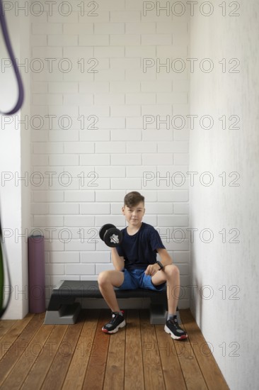 A young boy sitting on an exercise bench lifts a dumbbell in a home gym setting. He is wearing casual workout attire and appears focused on building strength and fitness