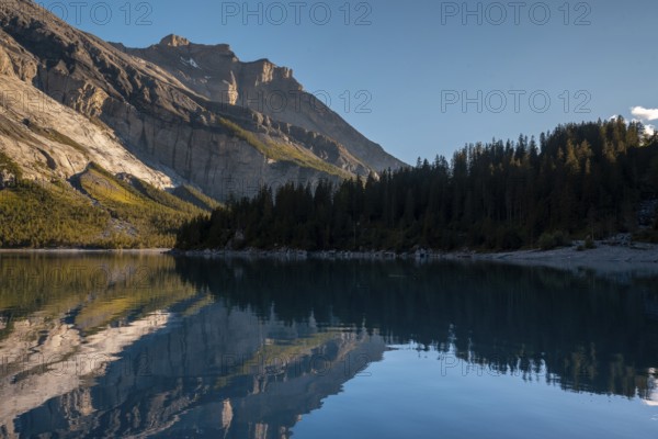 Tranquil lake Oachinensee in Switzerland reflects the majestic surrounding mountains and lush forests under a clear blue sky, offering a serene and picturesque landscape