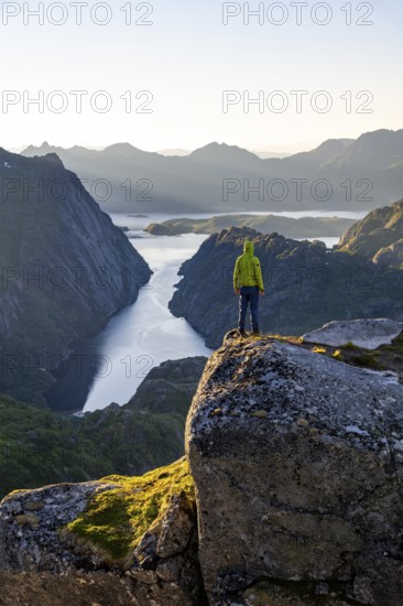 Mountaineer, young man standing on rocks and looking into the distance, at sunrise, fjord Trollfjord and mountains, hike to Trollfjord Hytta, at Trollfjord and Raftsund, Lofoten, Nordland, Norway