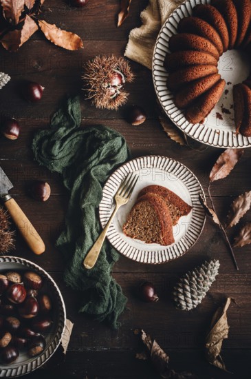 A beautifully styled chestnut cake on a ceramic plate, surrounded by whole chestnuts, leaves, and rustic decor, presenting a warm autumnal setting for a cozy treat