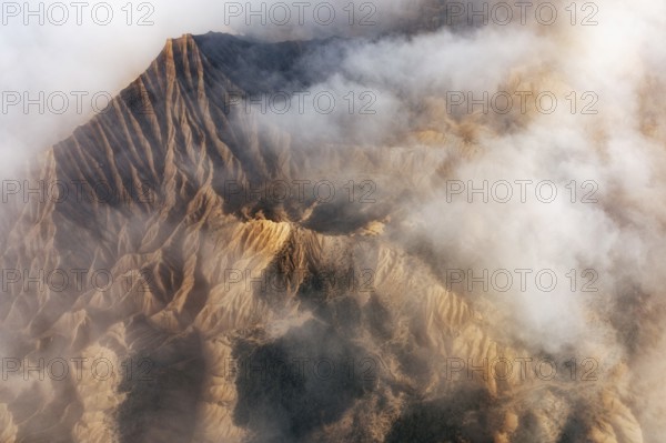 Aerial view of rugged mountain peaks partially covered by mist, creating a dramatic and serene natural landscape The sunlight enhances the textures of the terrain