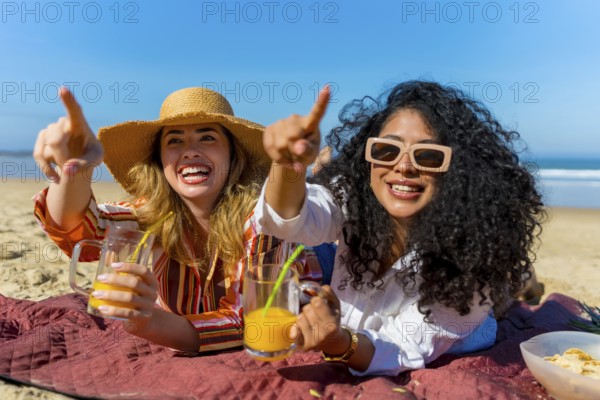 Two cheerful young women lying on a beach blanket, pointing into the distance with excitement while holding refreshing drinks, finding joy during their summer vacation