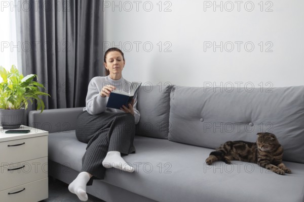 A woman reads a book on a gray couch in a cozy living room with a relaxed cat nearby. The setting features modern decor elements and a potted plant