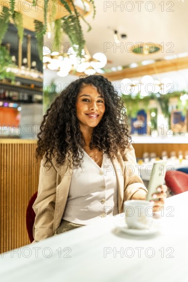Vertical photo of a happy latin woman using phone while drinking coffee in a modern cafeteria