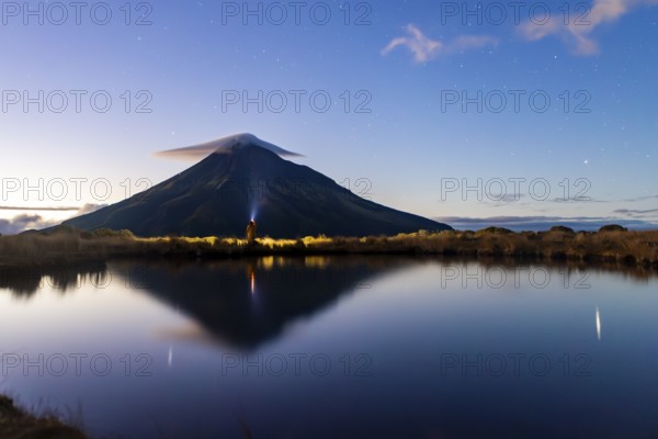 Back view of unrecognizable person looking at Mount Taranaki's reflection in a tranquil lake. The mountain, in Nueva Zelanda, is framed by a starry twilight sky