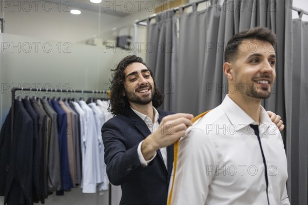 A tailor measures a smiling client suit in a modern suit store. The fitting room features various suits hanging, highlighting the personalized tailoring service