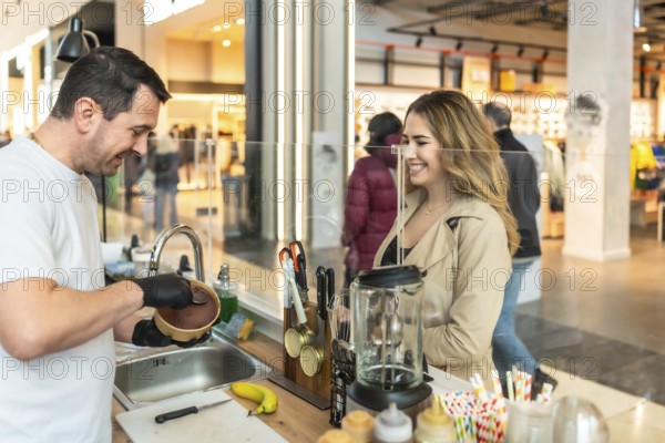 Man in apron serves a bowl of ice cream to a smiling young woman at a mall food stand counter, cheerful vendor customer interaction in a modern indoor shopping setting