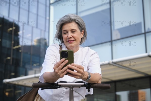 A mature woman with gray hair smiles as she uses a smartphone while standing with an electric scooter in an urban setting, illustrating modern lifestyle and technology
